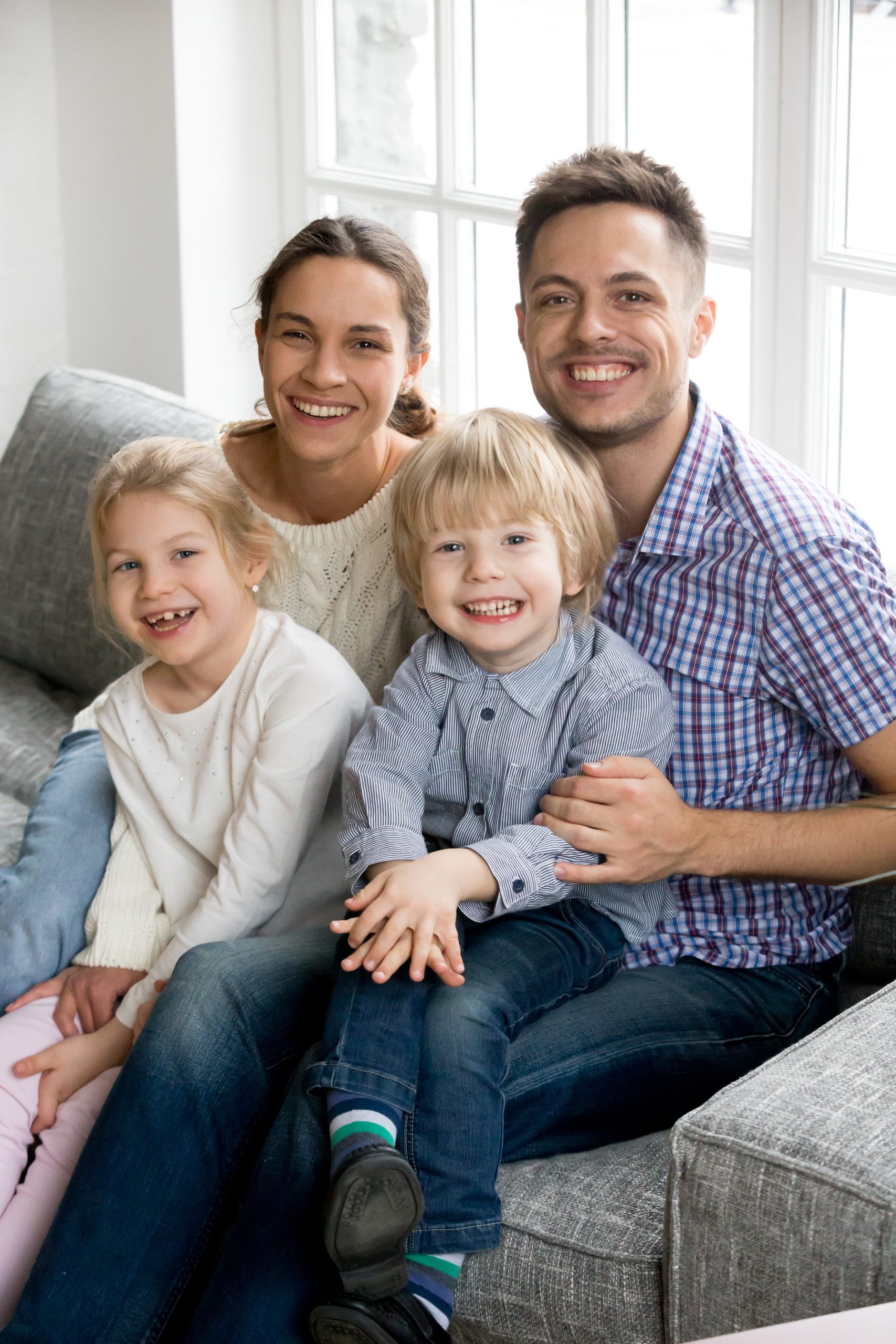 New happy parents for two adopted kids siblings concept, smiling couple posing with daughter and son sitting on sofa together, cheerful family bonding at home looking at camera, vertical portrait