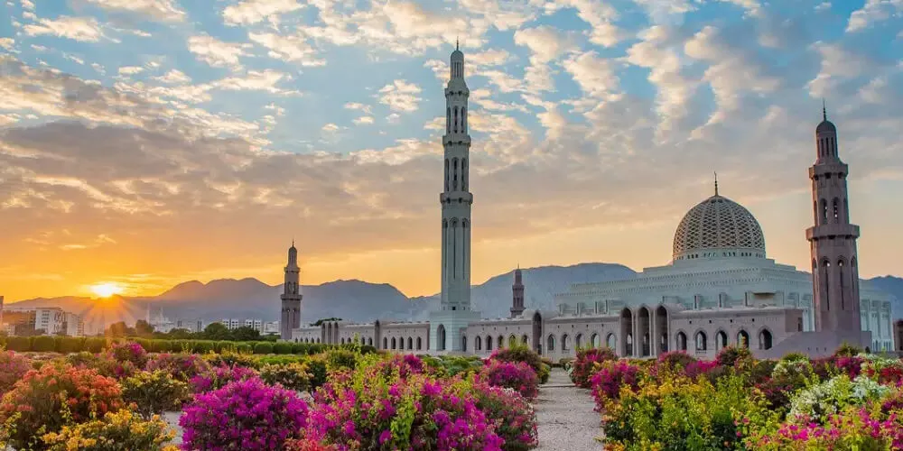 تصویر یک جای مذهبی در کشور عمان است که گنبد دارد و جلوش پر از گلهای صورتی و یک آسمان آبی پر از ابرهای زیبا دارد. بهترین سایت کاریابی در عمان